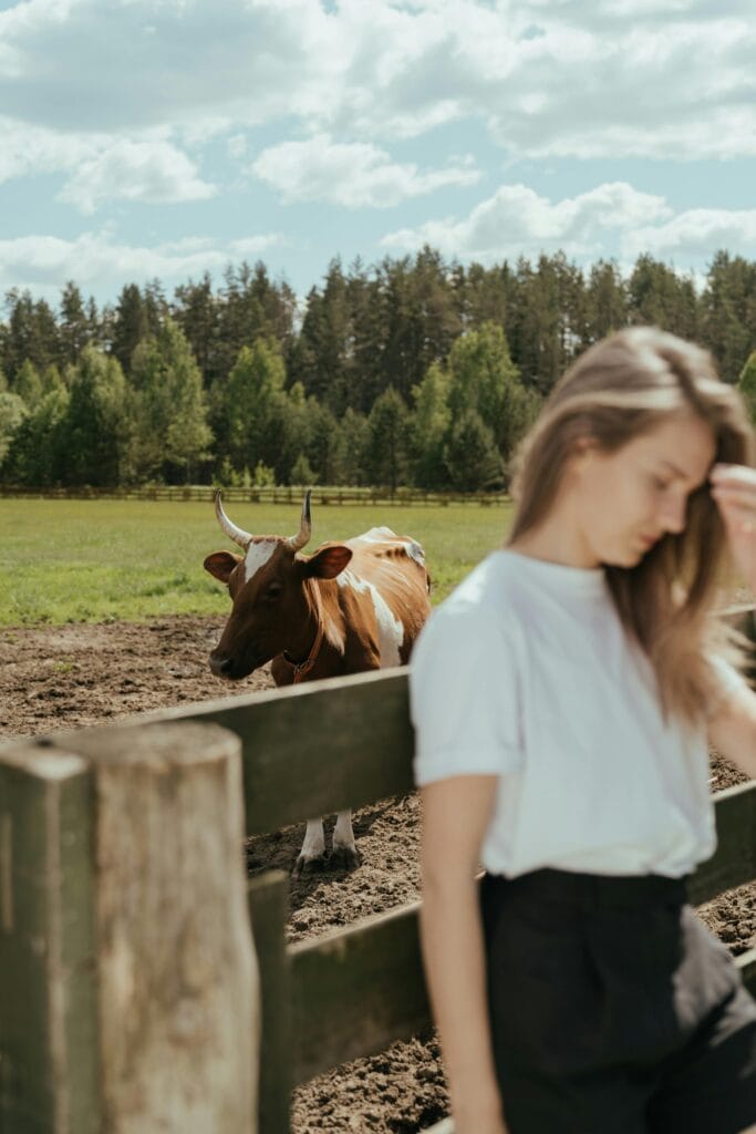 Woman in a farm environment standing near a fence, with a cow in pasture.